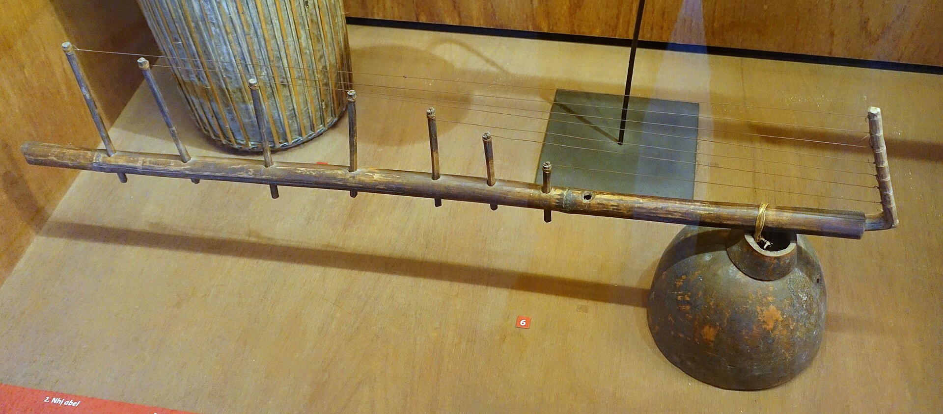 A long bamboo tube zither with pegs along it and a gourd resonator at one end, lying on a museum stand