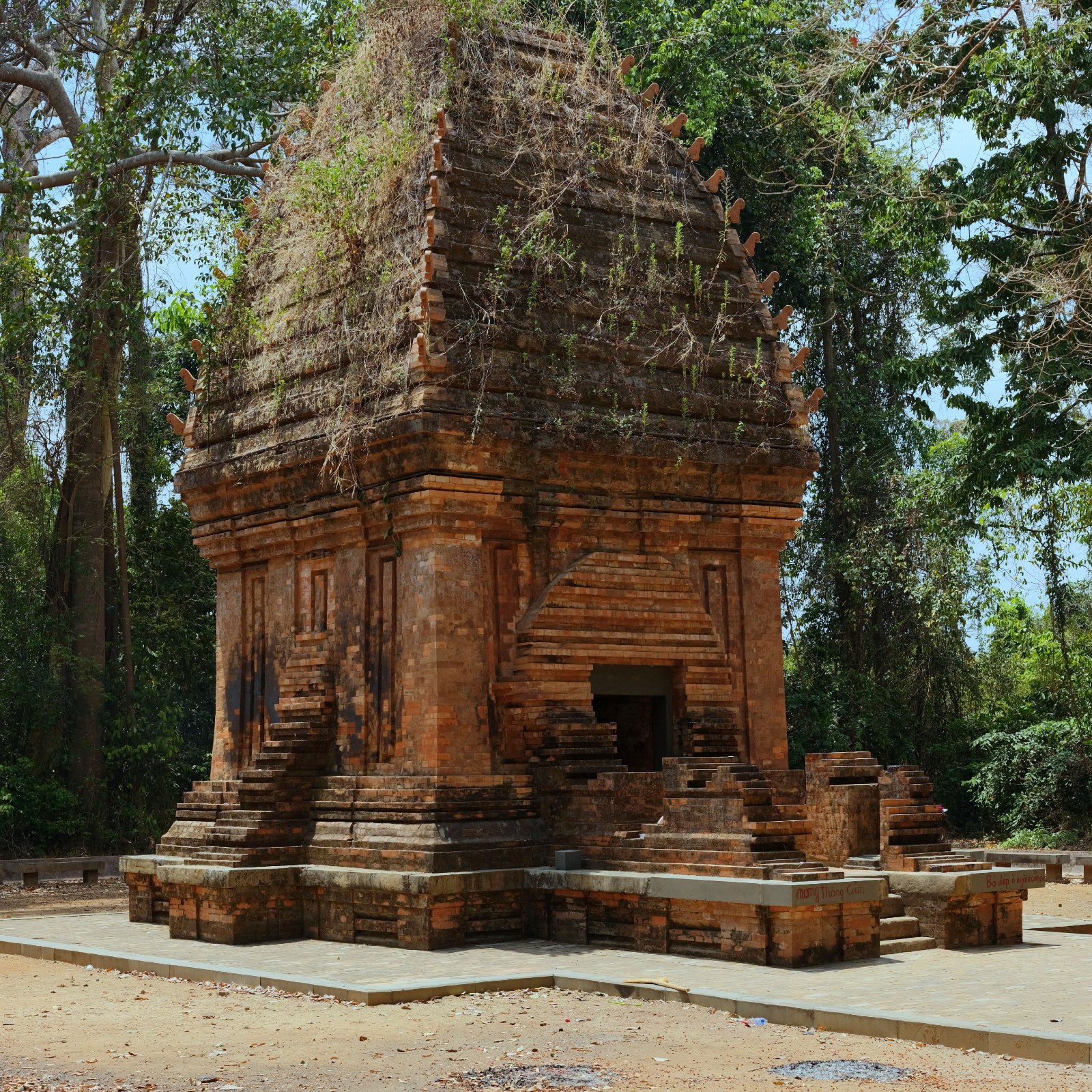 A brick Cham tower with a pointed roof standing among tall trees