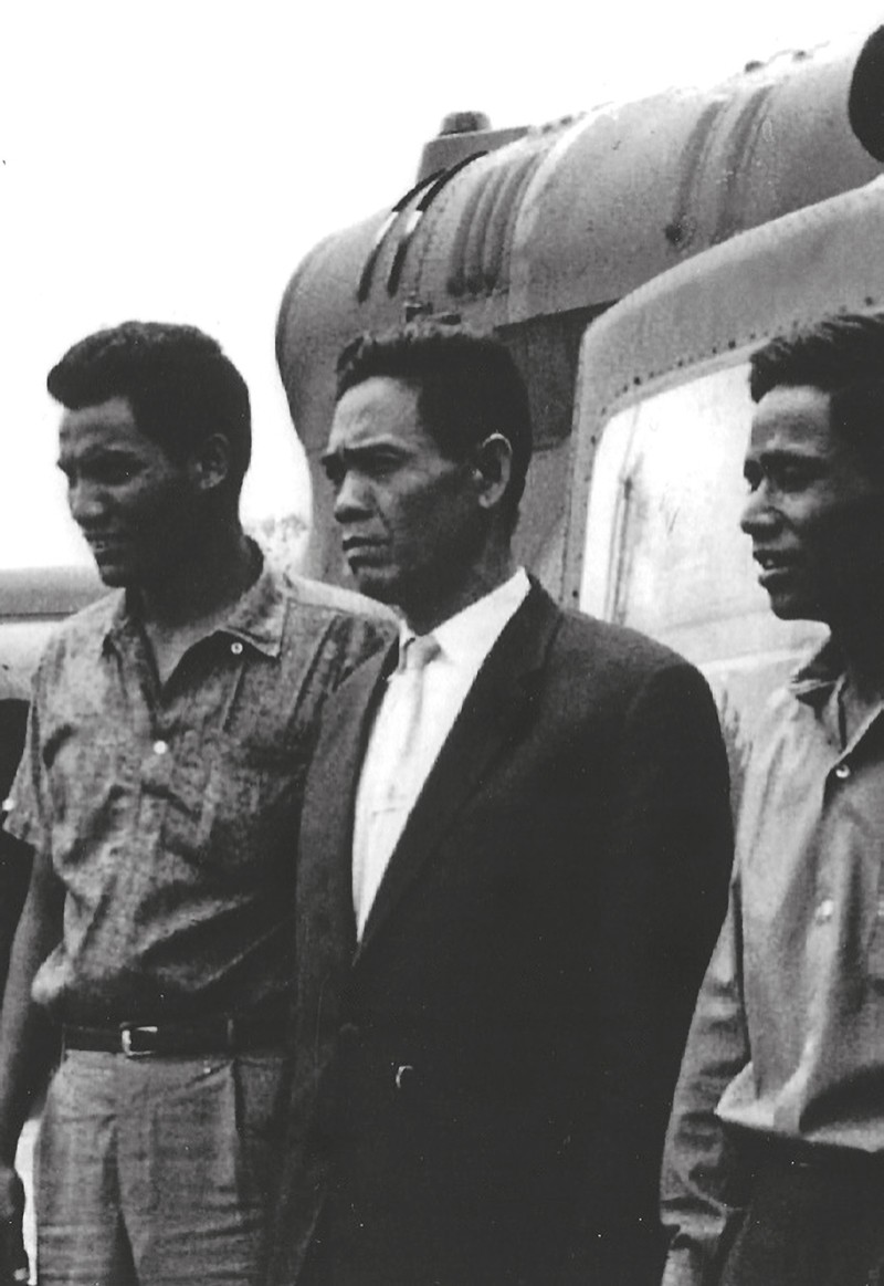 Three men standing beside an aircraft, the man in the middle in a dark suit and tie