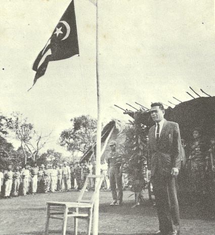 Y Bham Enuol in a suit standing at a flagpole, the flag flying above him, troops behind