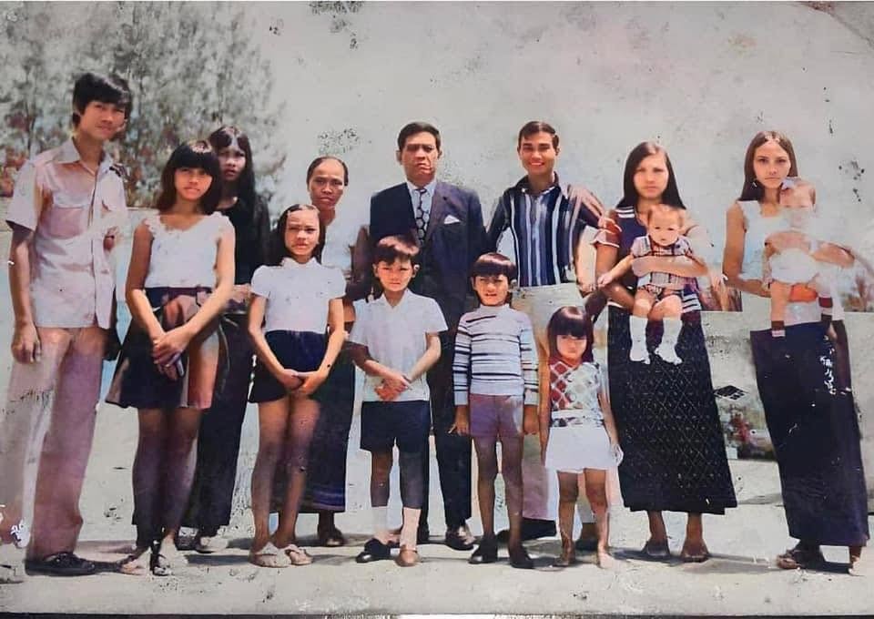 A family group of fifteen people photographed outdoors against a wall, a man in a dark suit at the center